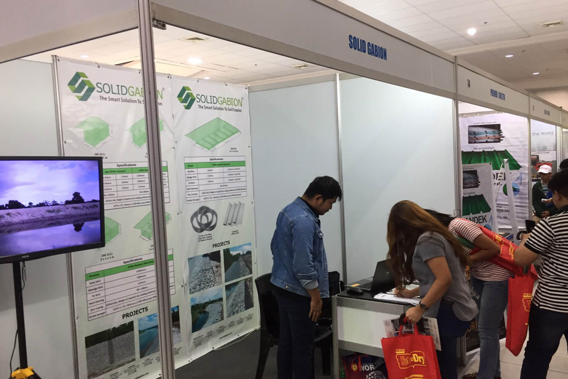 People at a Solid Gabion trade show booth with product information displays and a video screen.