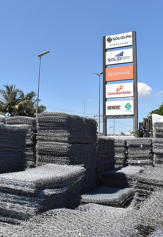 Stacked gabion mesh wire with a sign listing solar energy companies in the background.