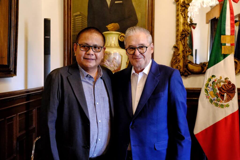 Two men in suits stand together, with the Mexican flag visible in the background.