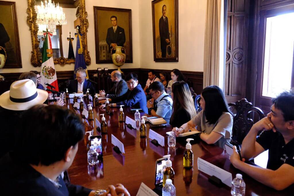 Group of people at a meeting seated around a long table with the Mexican flag visible.